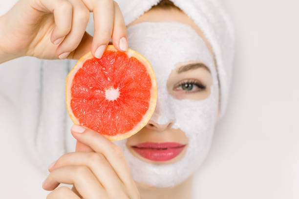 Top view portrait of charming young smiling woman with towel on head, facial mud clay mask, holding grapefruit slice hiding her eye, lying on white background. Close up, focus on grapefruit.